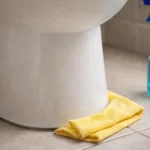 Pano amarelo de limpeza úmido e borrifador ao lado da base de um vaso sanitário no chão do banheiro.